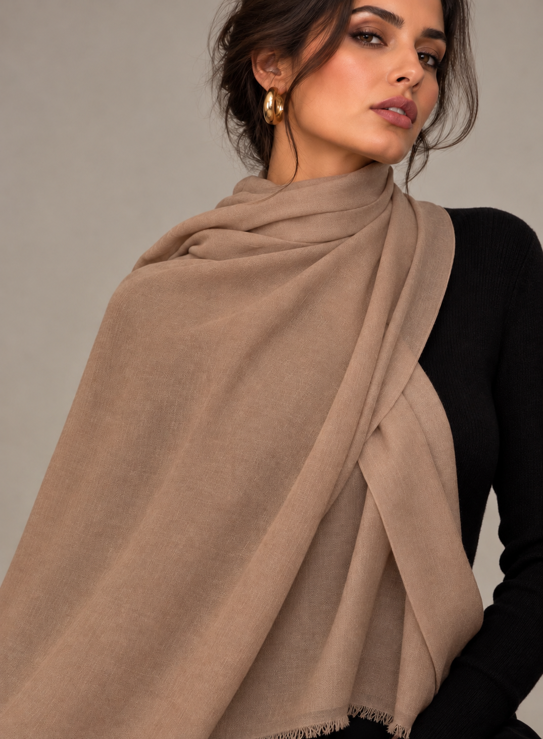 A woman wearing a soft taupe-beige pashmina shawl draped elegantly over her shoulders, paired with a black long-sleeve top and gold hoop earrings, against a neutral gray background.