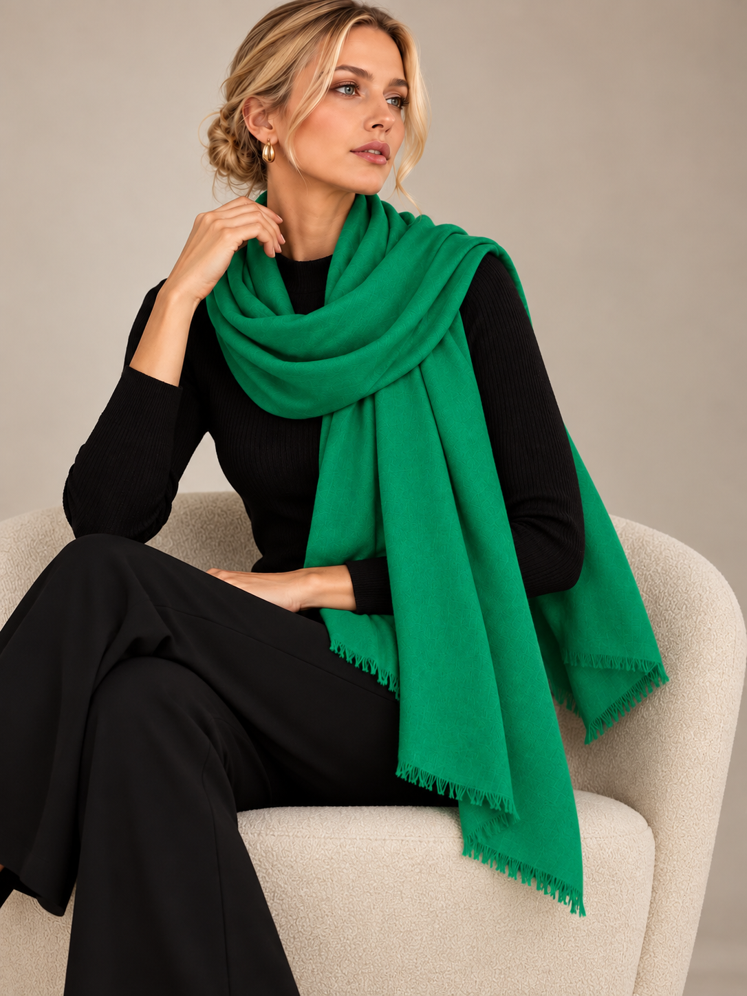 A woman seated in a light beige armchair wearing a bright green shawl over a black outfit, photographed against a soft neutral studio background.