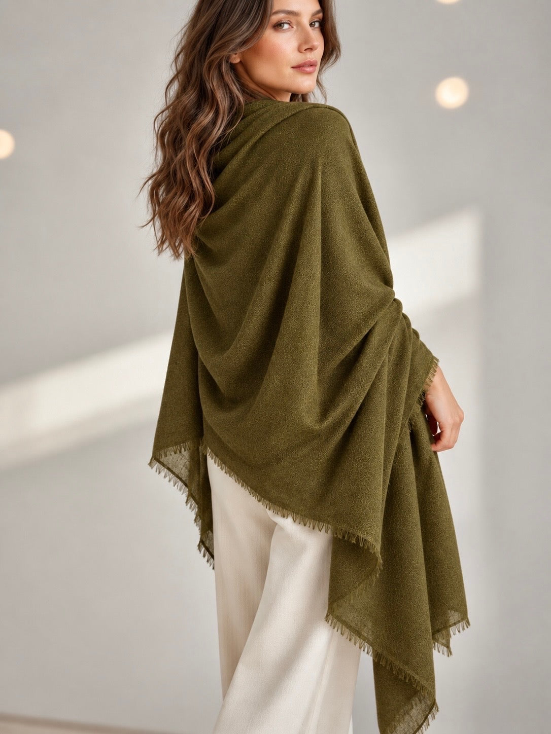 A woman wearing a draped olive green shawl with fringed edges over cream-colored pants, photographed in a softly lit indoor setting.