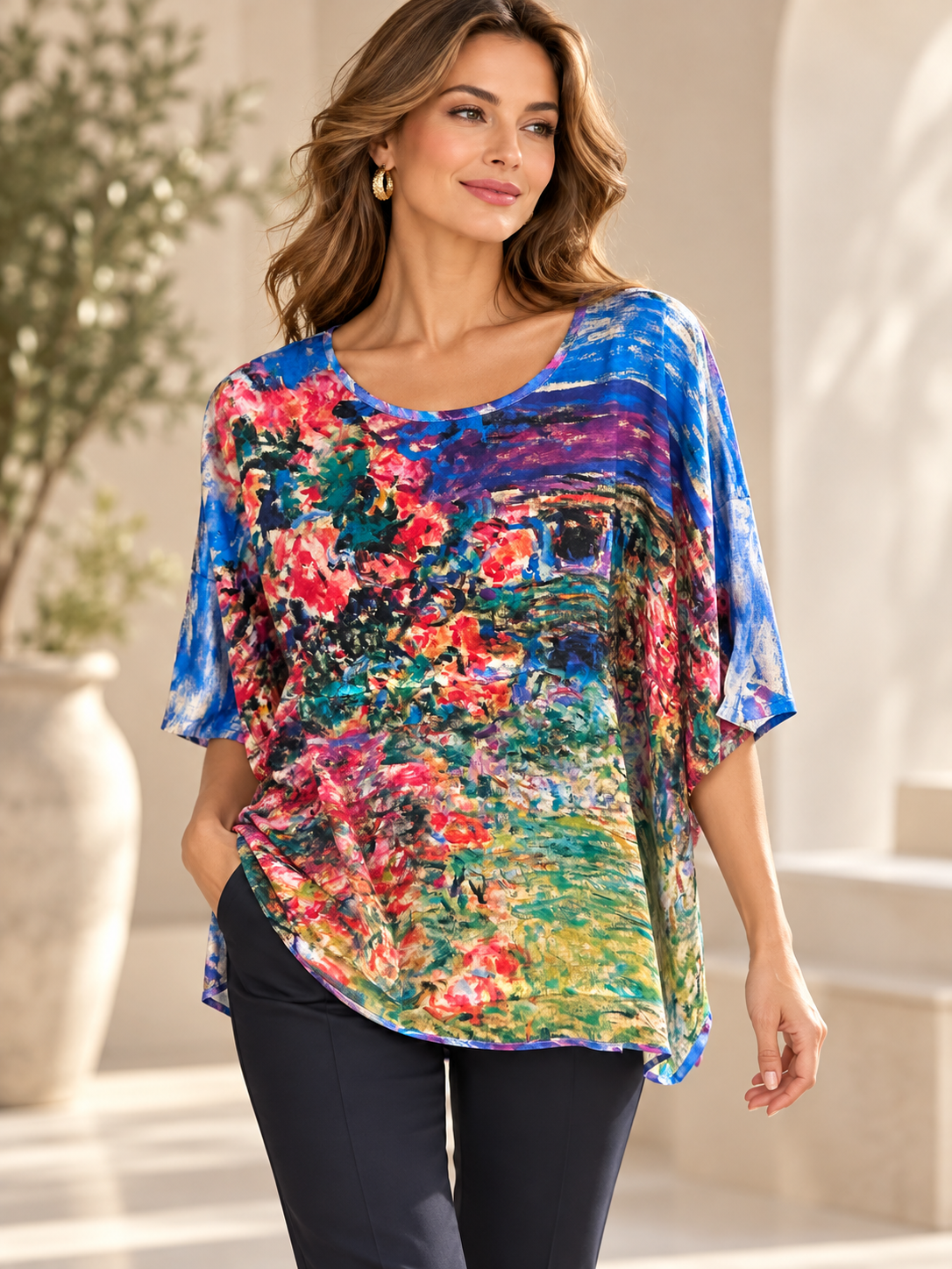 A woman wearing a vibrant, multicolored short-sleeve tunic with a painterly floral and landscape print, paired with dark pants, standing in a bright, sunlit indoor setting.