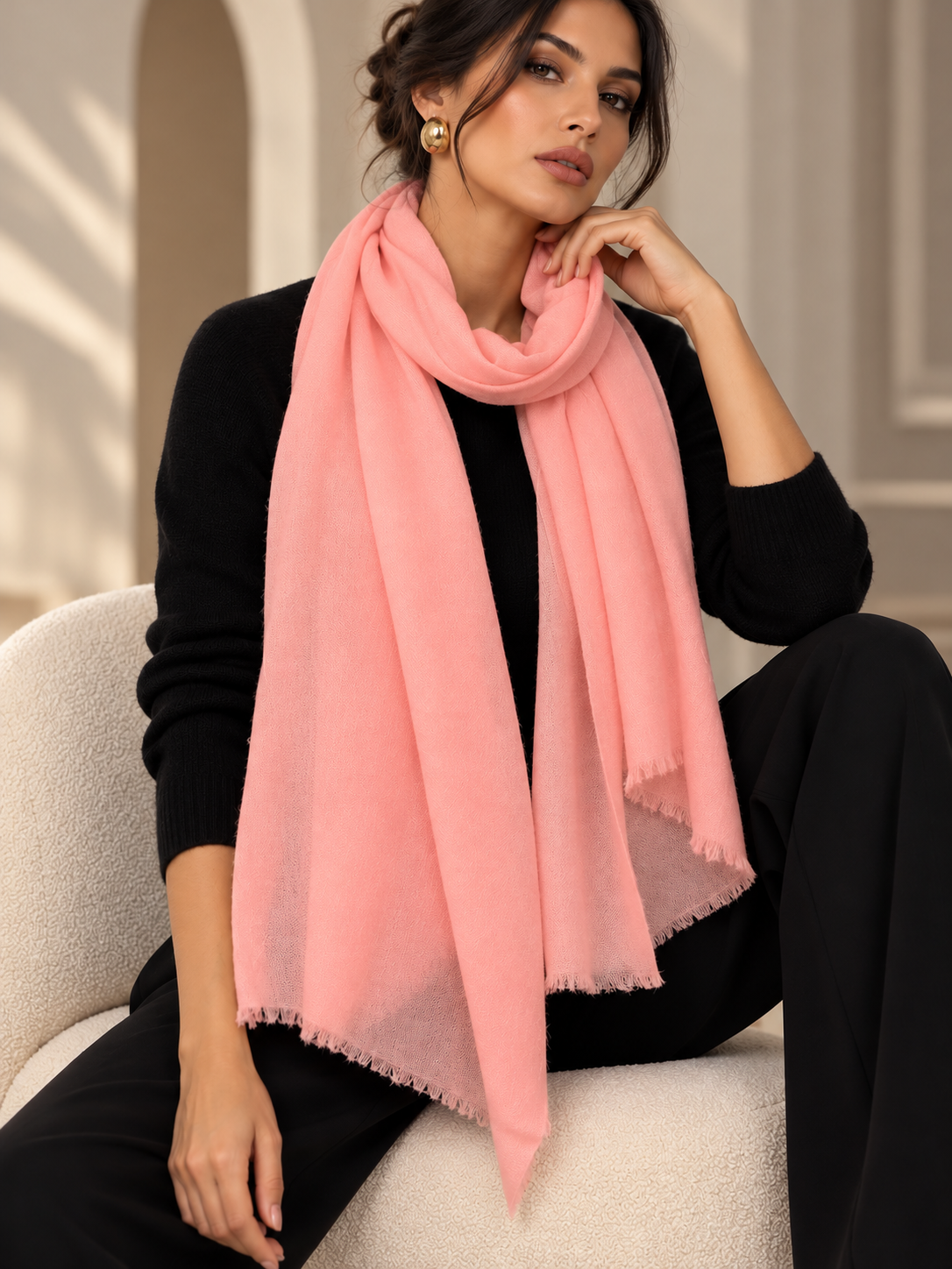 A woman wearing a soft pink scarf draped over her shoulders, styled with a black knit top and black pants while sitting on a cream-colored armchair.