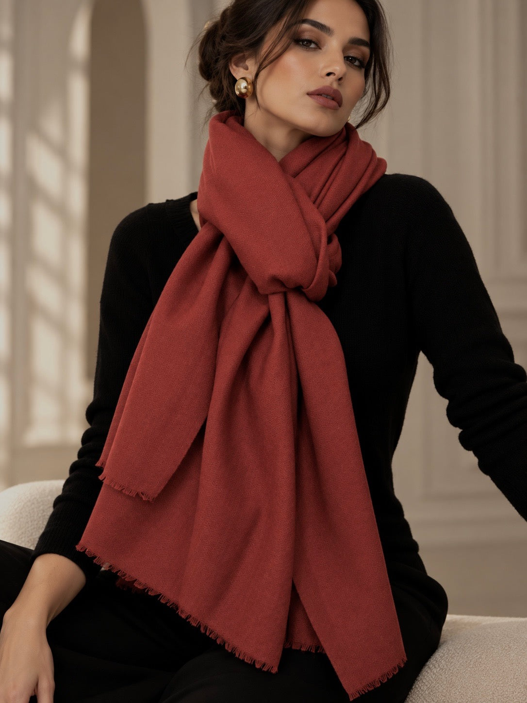 A woman wearing a rust-red woven scarf with fringed edges, styled over a black long-sleeve top, sitting in a softly lit indoor setting.