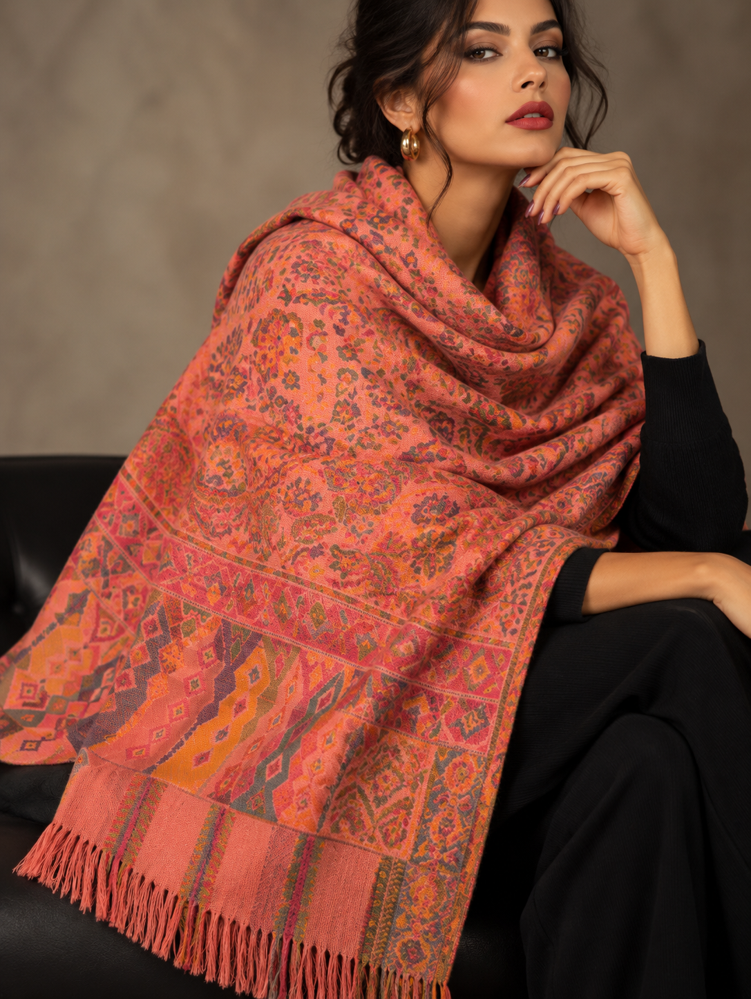 A woman wearing a large patterned shawl in coral, orange, and pink tones with geometric borders and fringe, styled over a black outfit against a neutral studio background.