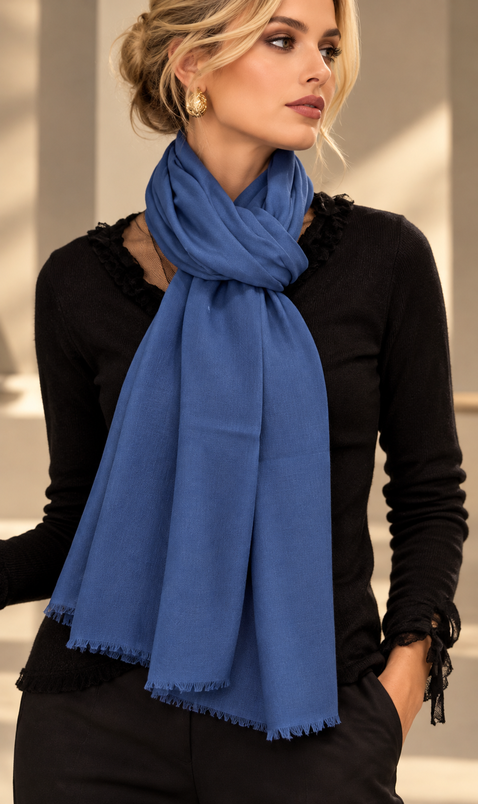 A woman wearing a soft blue scarf with fringed edges, styled over a black long-sleeve top in a bright indoor setting.