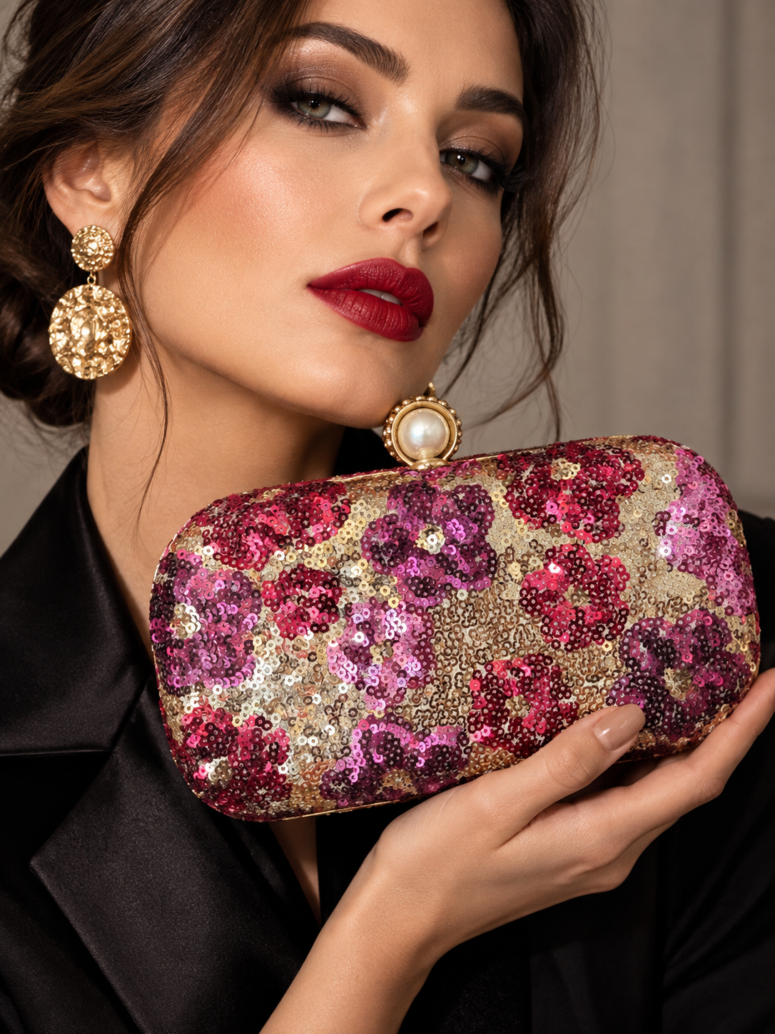 A close-up of a woman holding a rectangular clutch bag covered in pink, magenta, and gold sequins, with matching statement earrings and a black satin top against a soft neutral background.