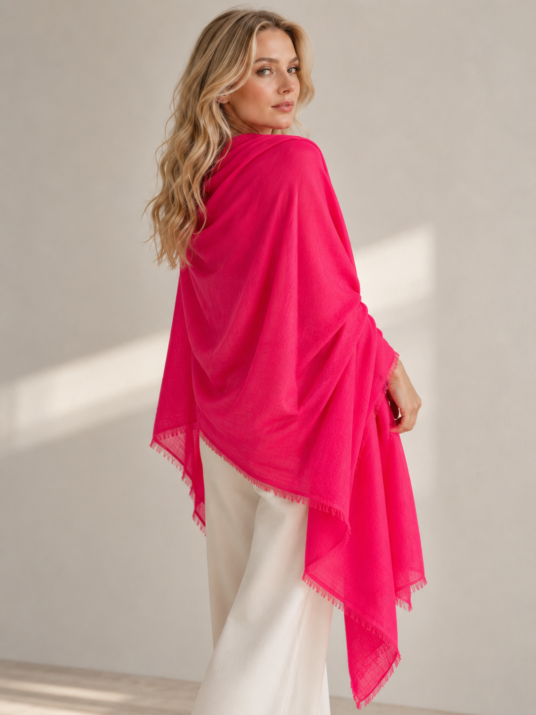 A woman wearing a bright pink shawl over white pants, photographed in a softly lit studio with a neutral beige background.