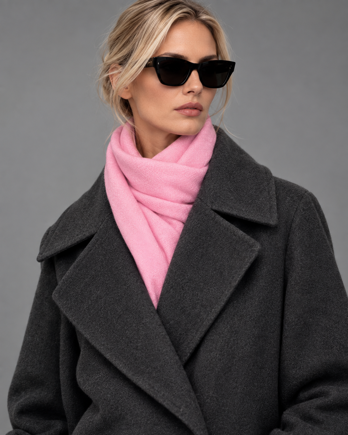A woman wearing a dark charcoal gray coat, a bright pink knit scarf, and black sunglasses, photographed against a solid gray studio background.