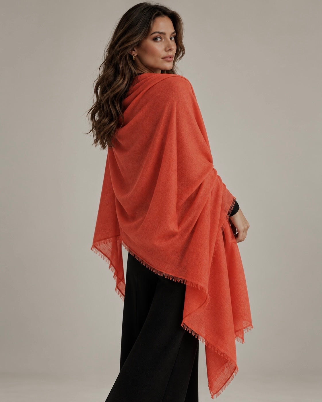 A woman wearing a bright orange-red shawl wrap with fringed edges over black pants, photographed against a plain light beige studio background.