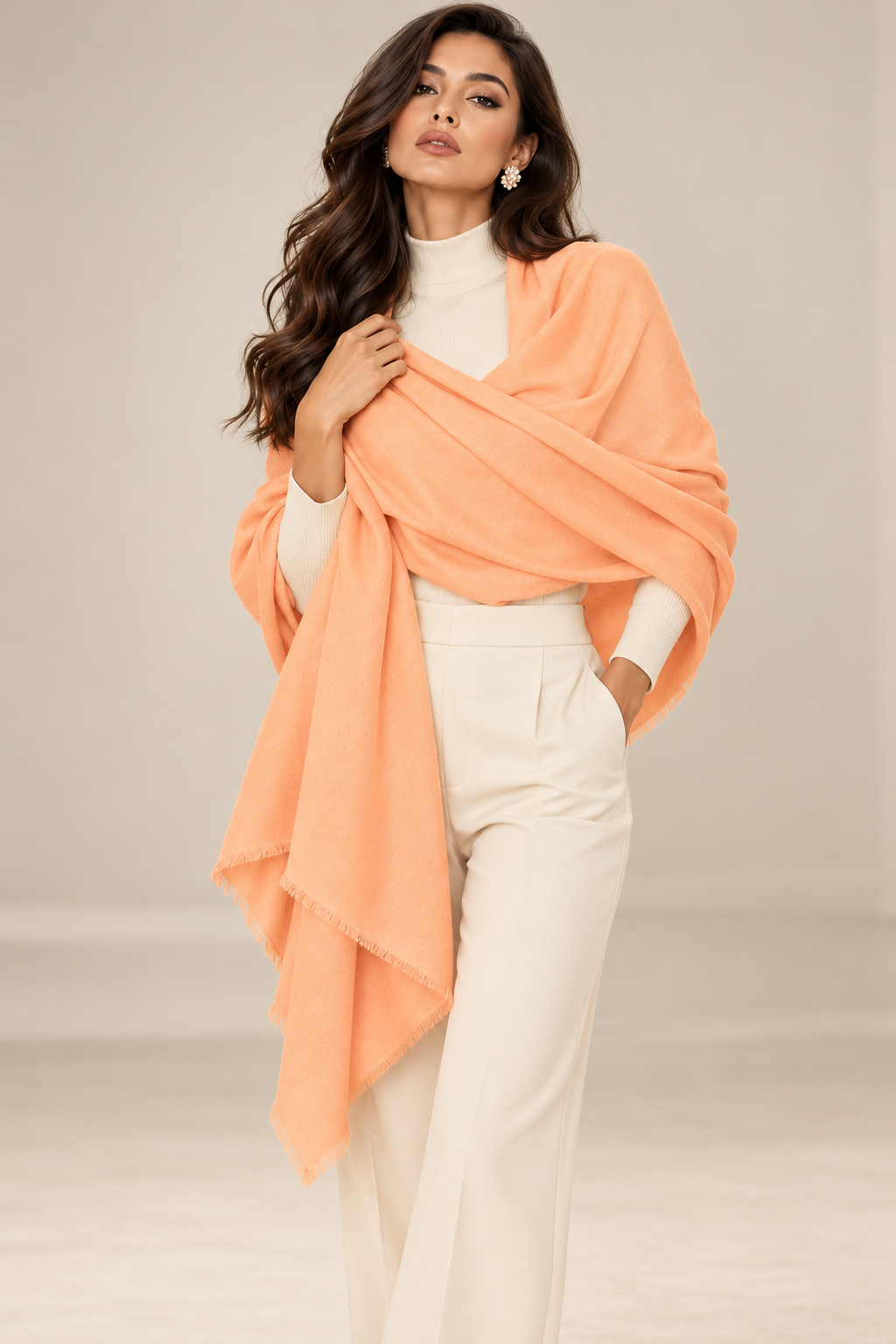A woman modeling a soft peach-orange shawl draped over a white mock-neck top and white wide-leg trousers, set against a neutral light gray studio background.