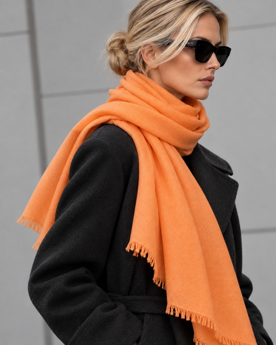A woman wearing a bright orange scarf and a black coat, photographed outdoors against a light gray wall.