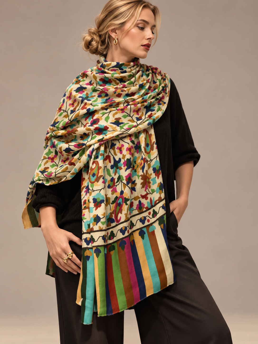 A woman models a multicolor printed scarf with a floral pattern and striped fringe, styled over a black outfit against a neutral studio background.