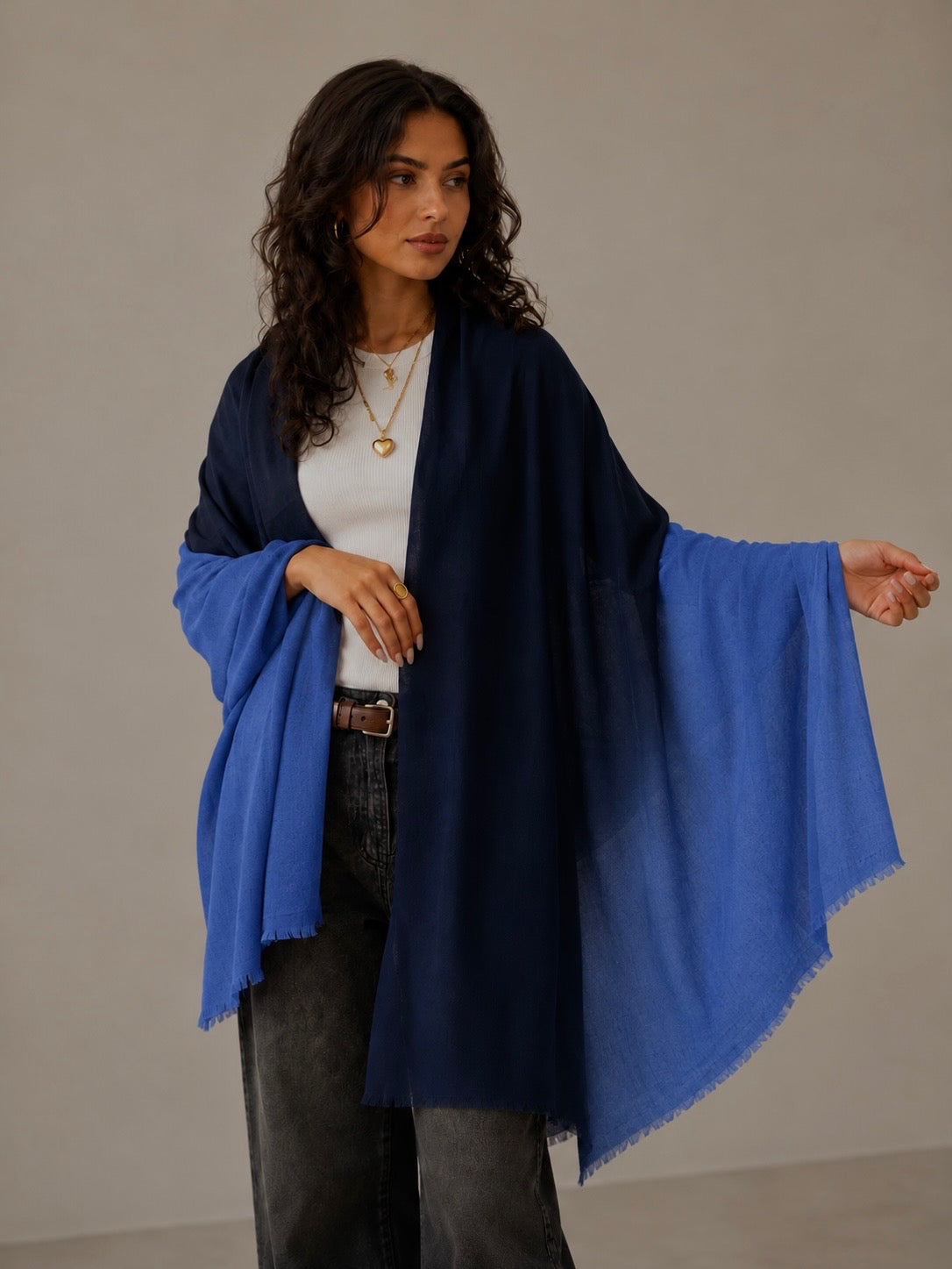 A woman modeling a navy blue shawl with a vibrant blue gradient transition effect, worn over a white top and dark gray jeans against a neutral beige background.