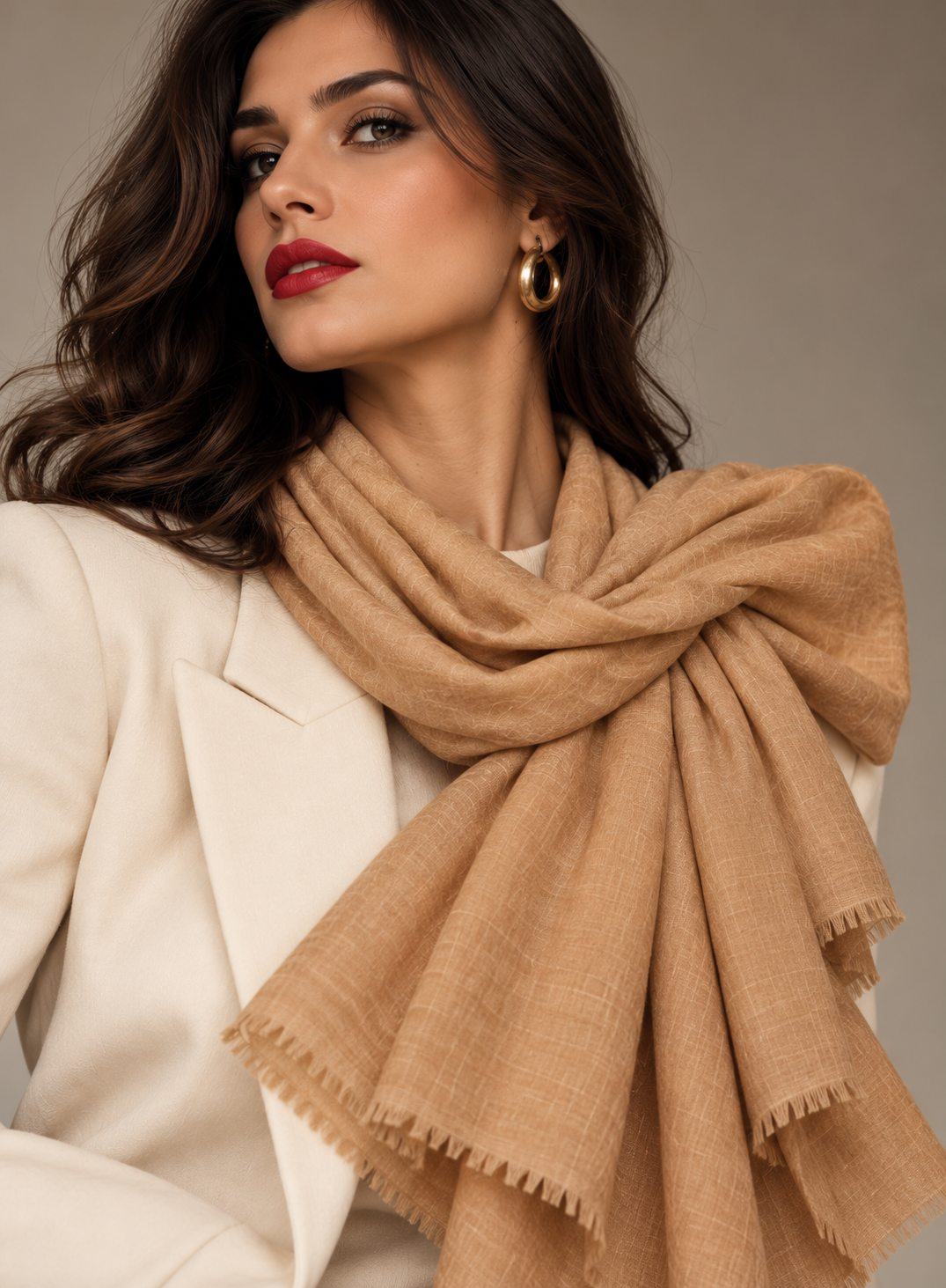 A woman wearing a cream-colored blazer and a tan woven scarf with fringed edges, styled with gold hoop earrings against a neutral gray background.