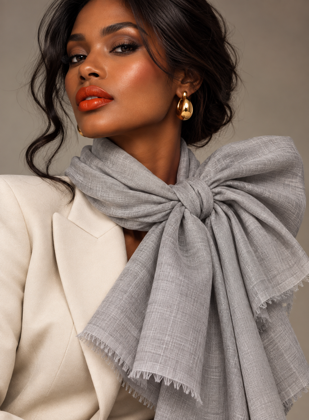 A woman wearing a light gray textured scarf tied in a large bow around her neck, paired with a cream blazer and gold teardrop earrings, against a neutral beige background.