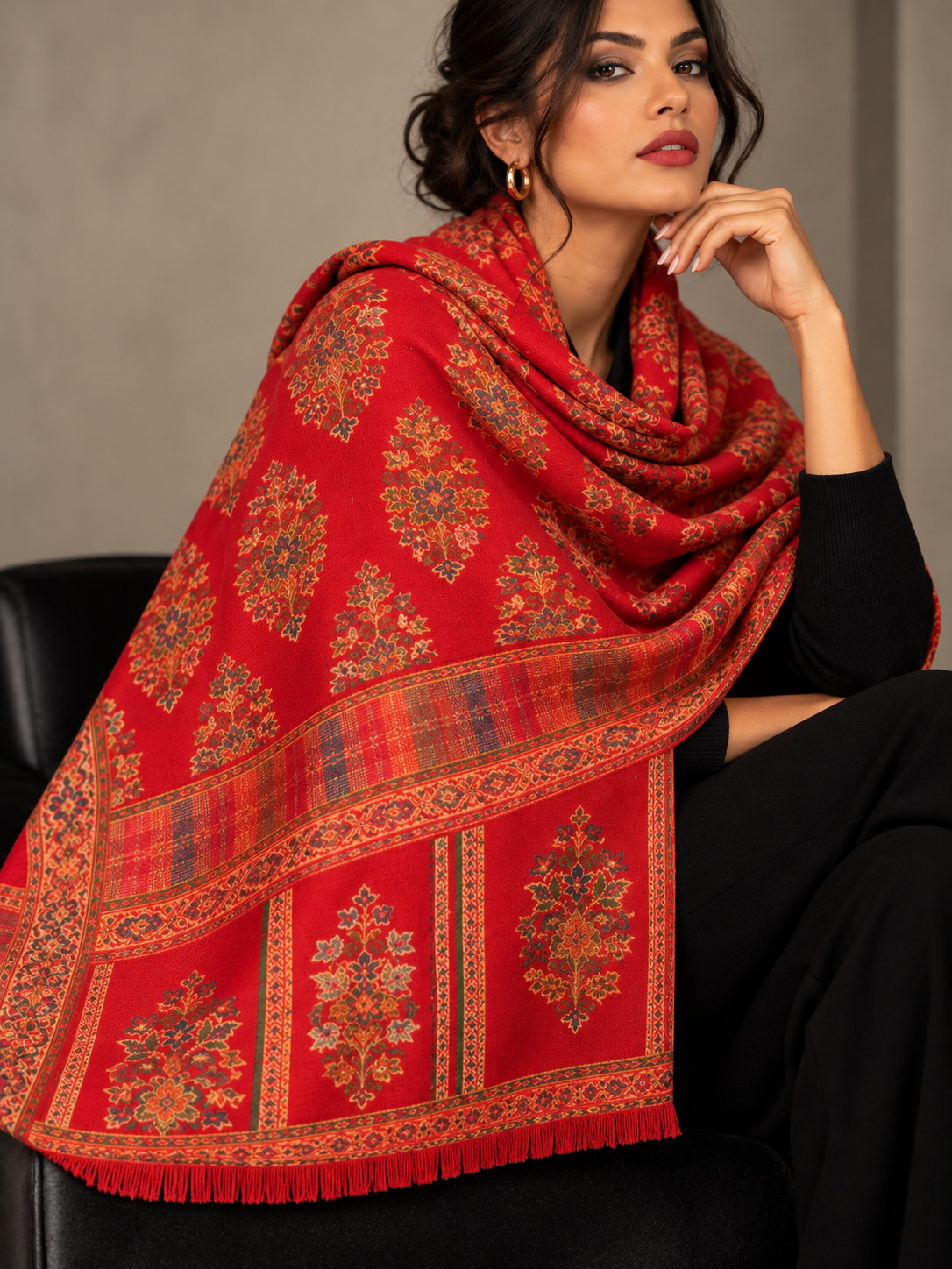A seated woman wearing a red shawl with gold-toned floral motifs and an ornate border over a black outfit.