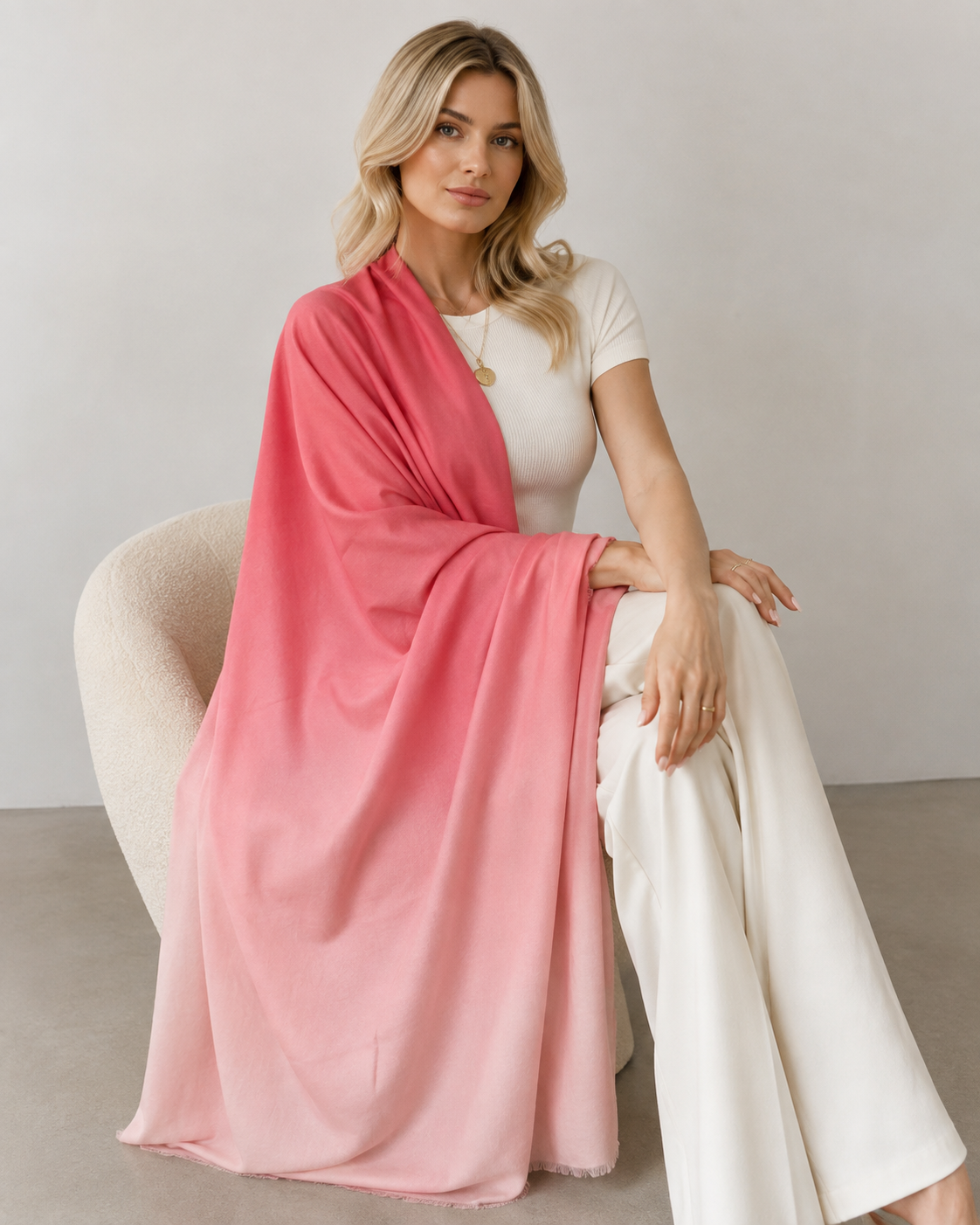 A woman sits on a cream-colored chair wearing a pink ombre gradient shawl draped over her shoulders, paired with a white top and white wide-leg pants, against a neutral studio background.