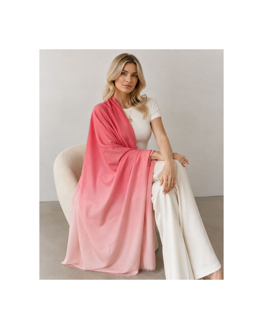 A woman sits on a cream-colored chair wearing a pink ombre gradient shawl draped over her shoulders, paired with a white top and white wide-leg pants, against a neutral studio background.