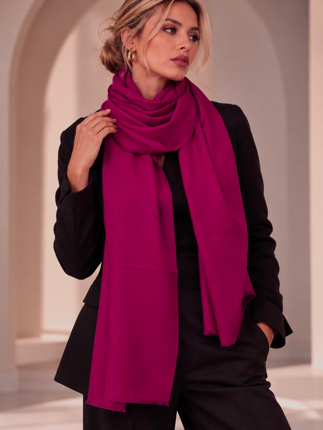 A woman wearing a bright magenta-pink lightweight scarf, styled with a black blazer and black trousers, standing in front of a soft beige arched background.
