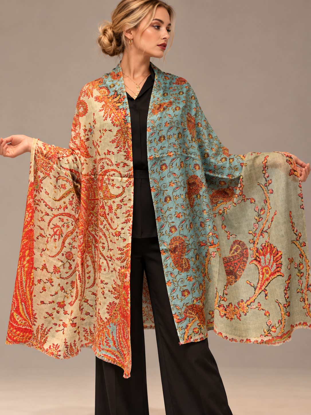 A woman models a lightweight open-front shawl or kimono-style wrap with wide sleeves, featuring a split-color paisley pattern in turquoise blue and warm rust orange on a beige base, over a black outfit against a neutral studio background.