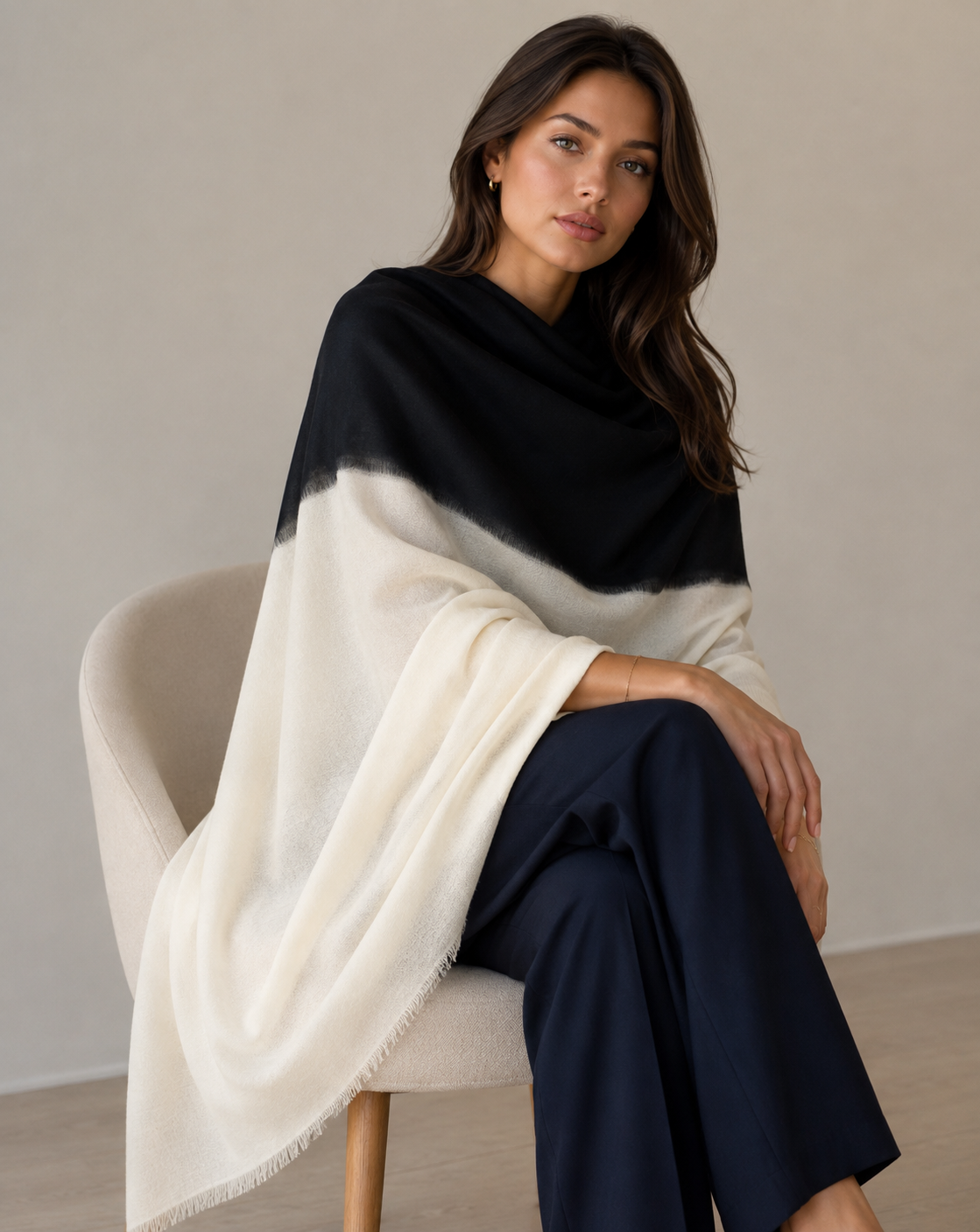 A woman seated on a light upholstered chair wearing a large color-blocked shawl or wrap in black and off-white, paired with dark navy pants, against a neutral indoor wall.