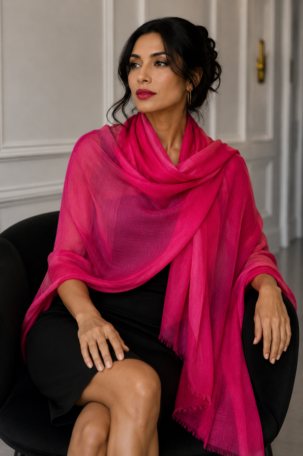 A woman seated in a black armchair wearing a bright pink handwoven cashmere shawl draped over her shoulders, paired with a black dress