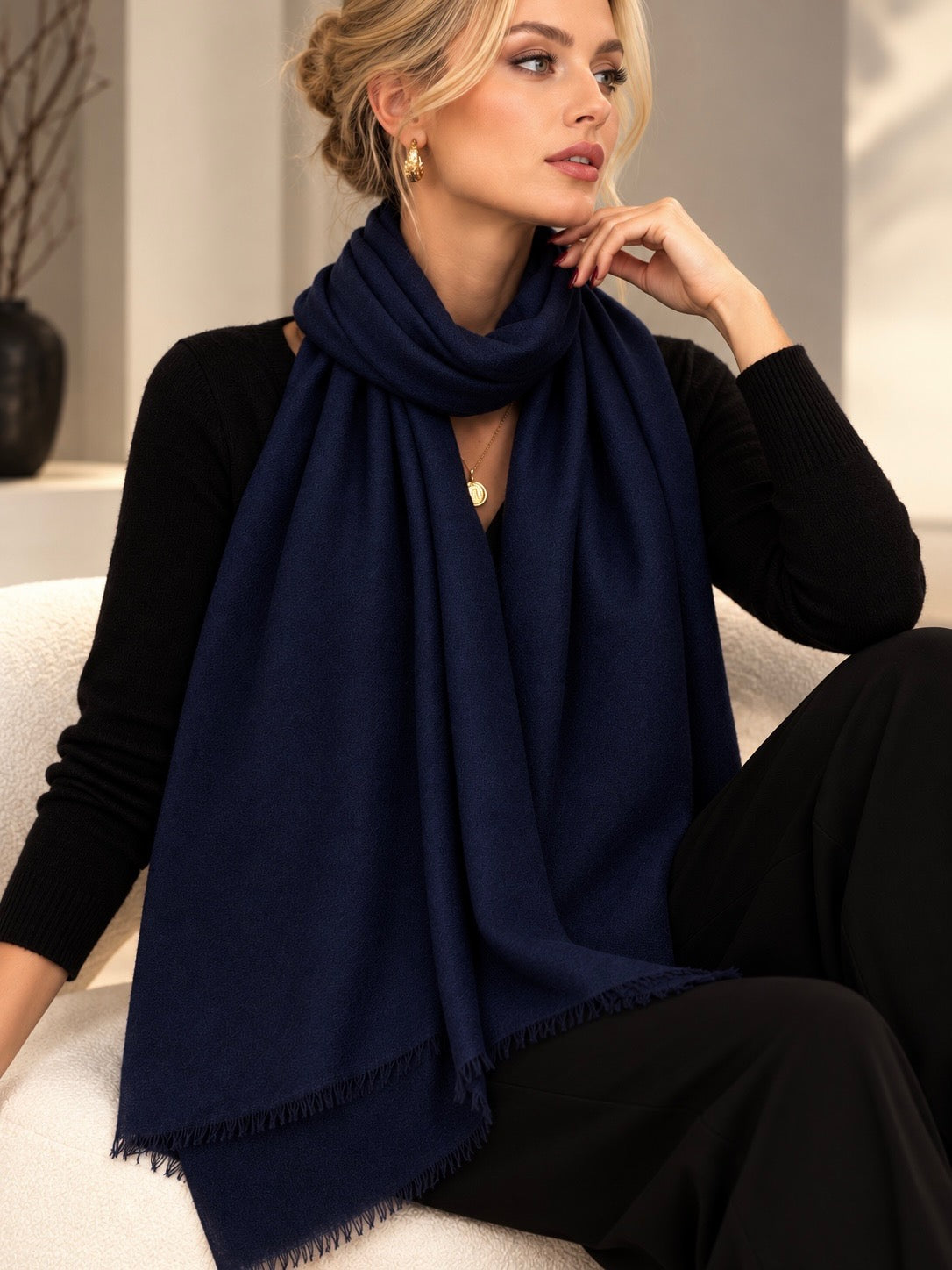 A woman wearing a navy blue scarf draped over her shoulders, paired with a black long-sleeve top and black pants, sitting on a white sofa in a softly lit indoor setting.