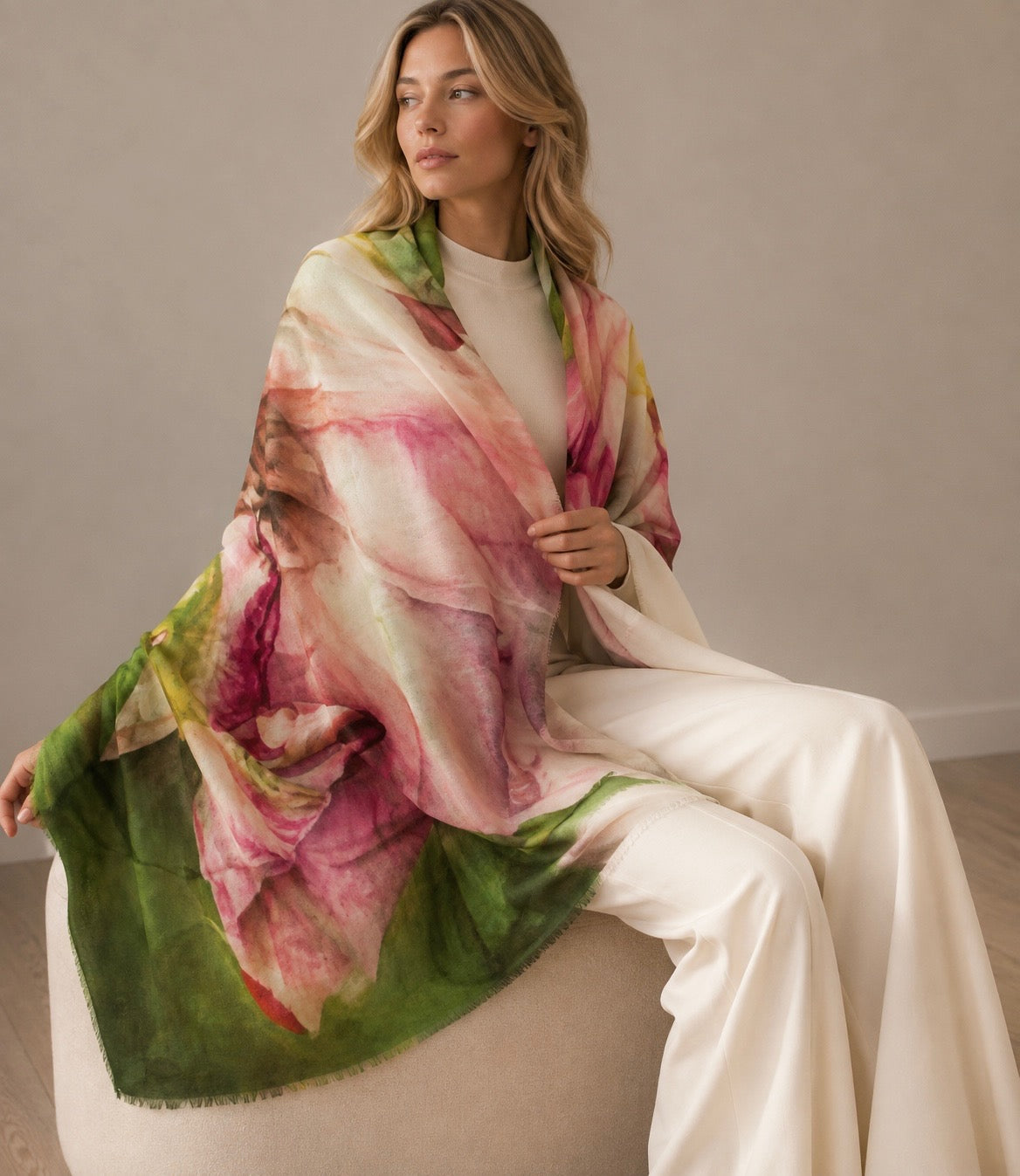 A woman sits on a round upholstered chair wearing a large pastel shawl with a watercolor-style floral print in pink, cream, green, and touches of yellow. The background is a clean, neutral beige studio setting.