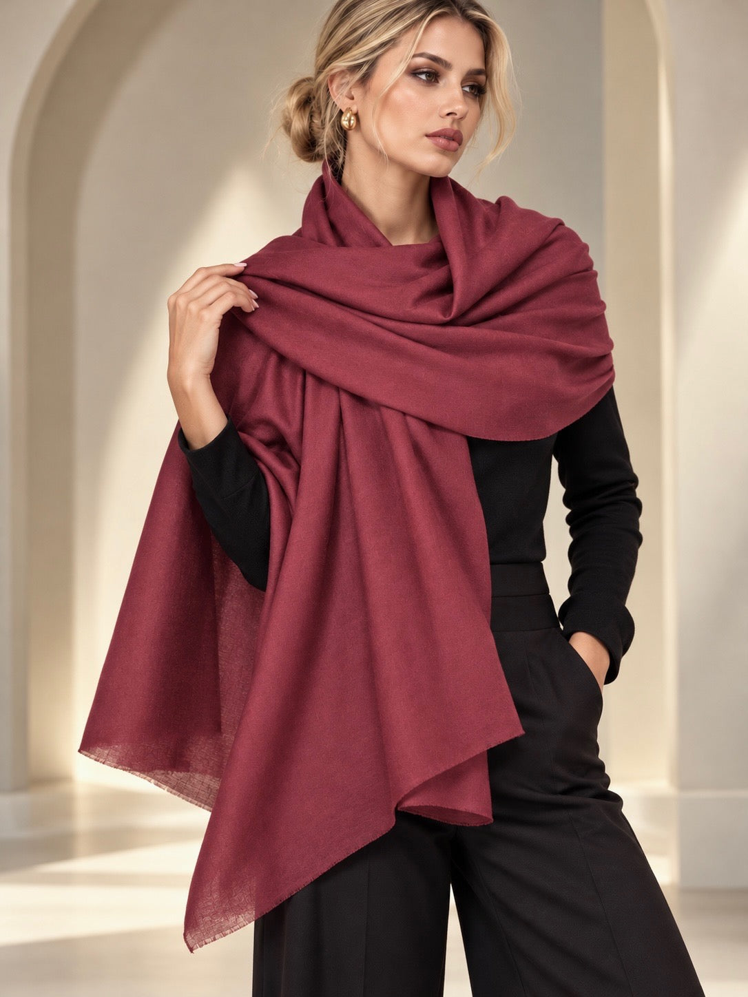 A woman models a lightweight, draped shawl in a deep burgundy or maroon shade, styled over a black outfit in a softly lit, elegant interior setting.