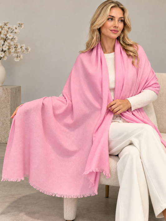 A woman seated on a light upholstered chair wearing a soft pink shawl over a white top and white wide-leg pants in a neutral indoor setting.