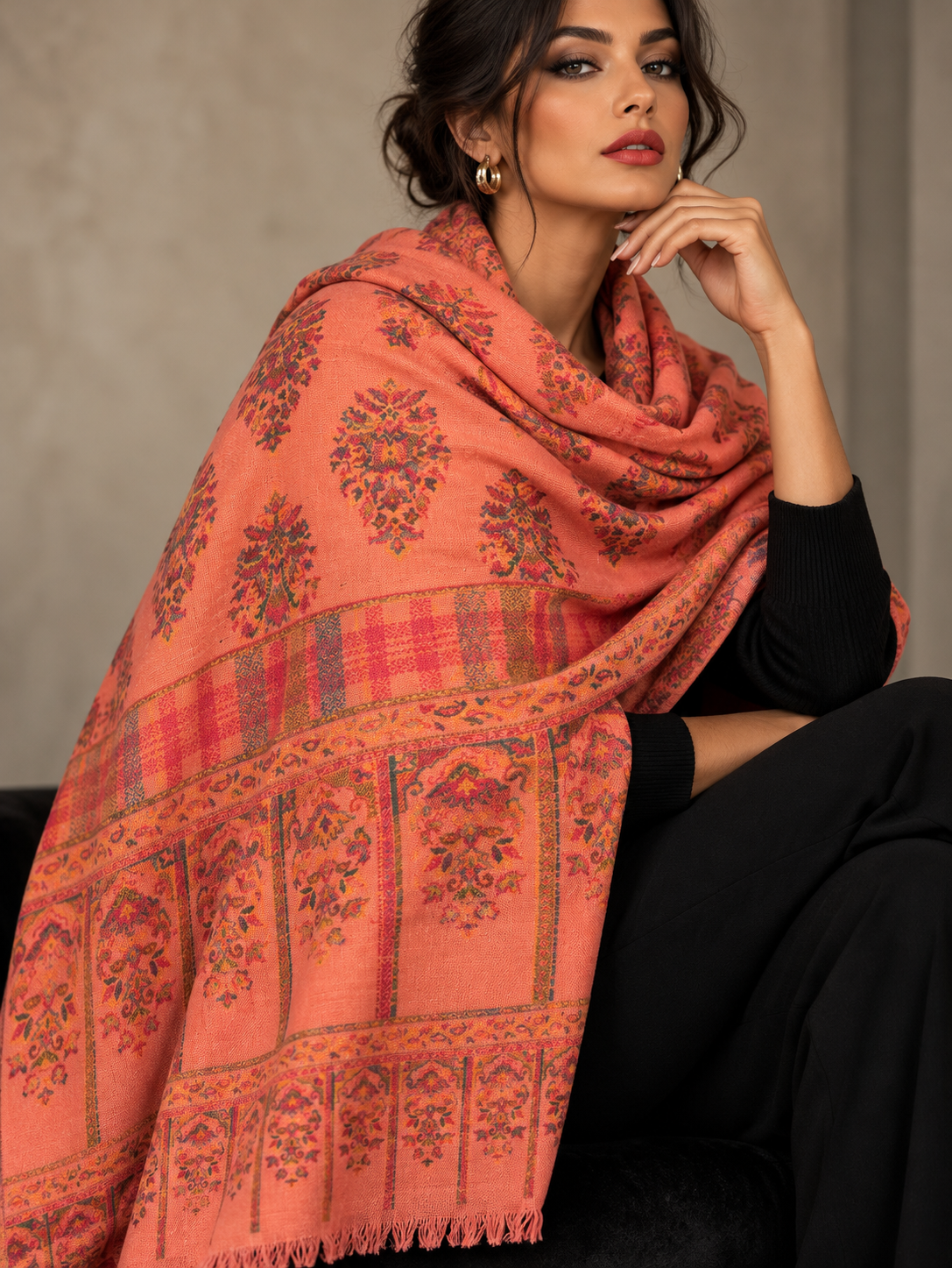 A woman wearing a large peach-pink shawl with intricate red, orange, and dark floral patterns and a plaid border, styled over a black long-sleeve top and black pants.