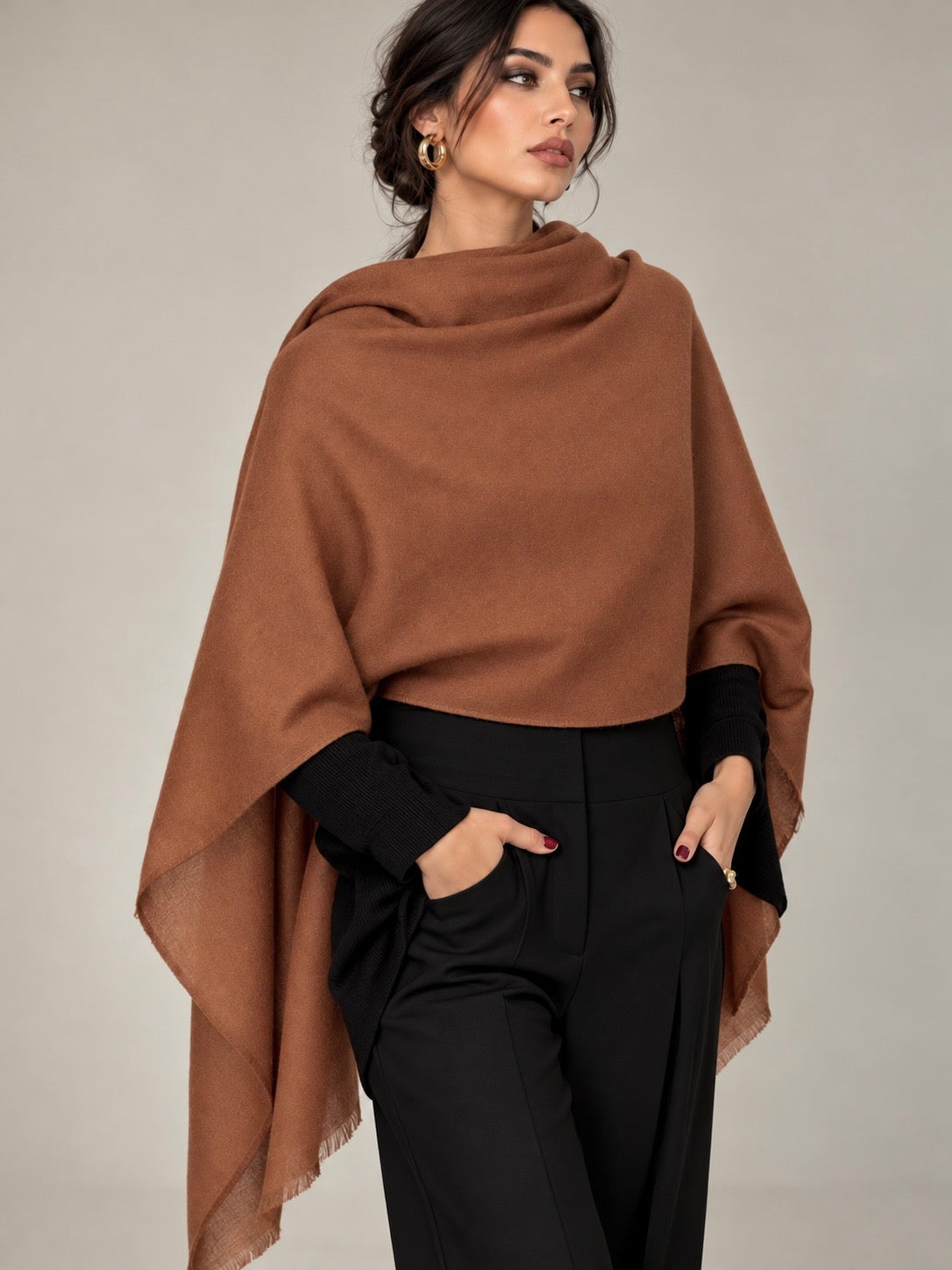 A woman models a draped camel brown shawl or poncho over a black long-sleeve top and black trousers, set against a plain light gray studio background.