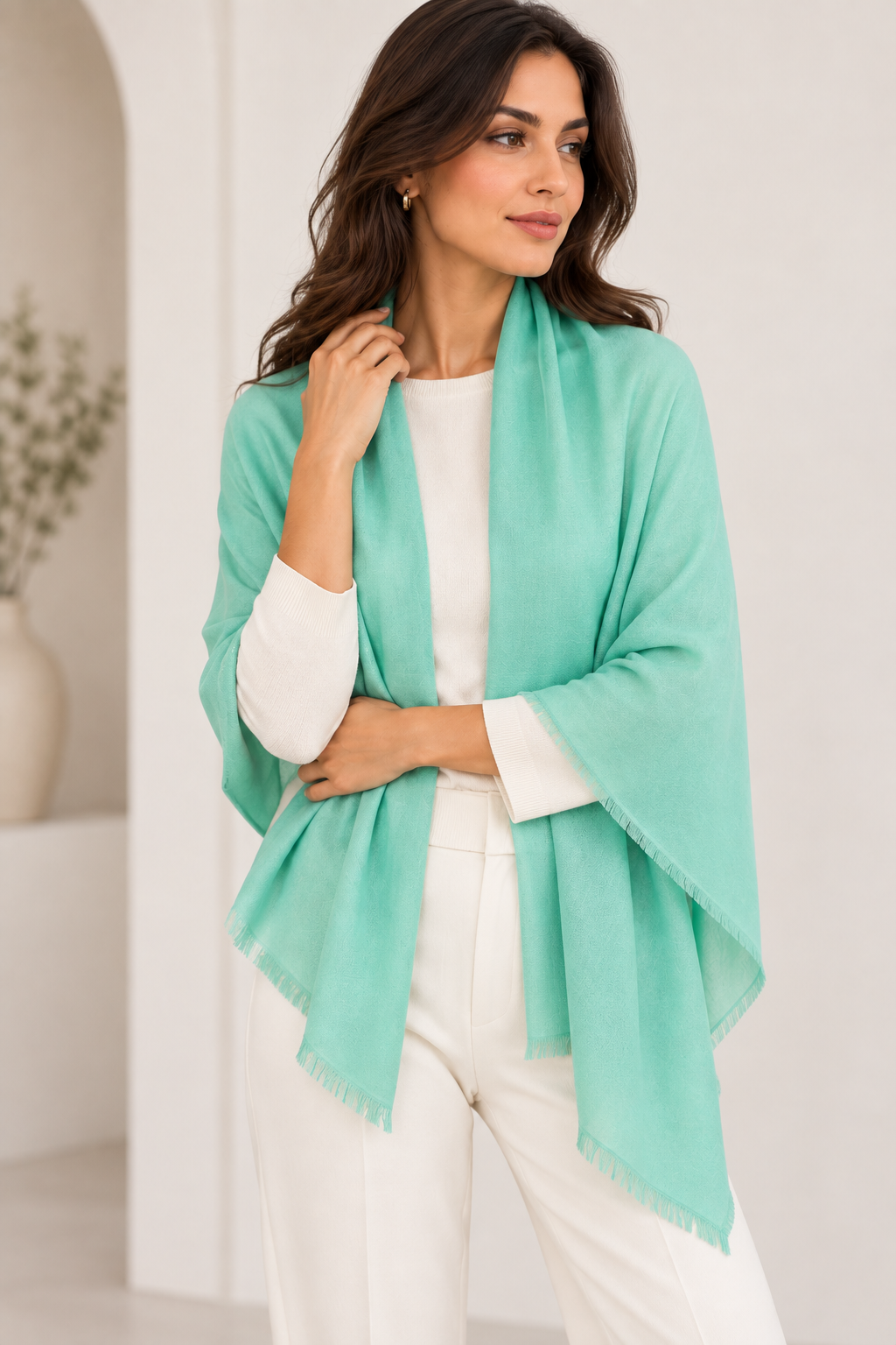 A woman modeling a light mint green shawl with fringed edges over a white top and white pants, standing in a bright, minimalist white interior setting.