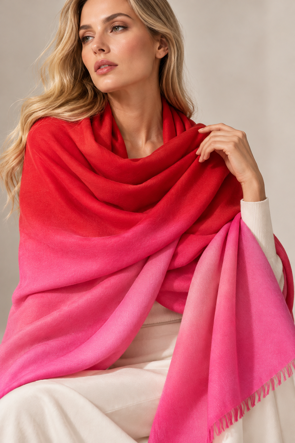 A woman draped in a large gradient shawl that transitions from bright red at the top to vivid pink at the bottom, posed against a soft beige studio background.