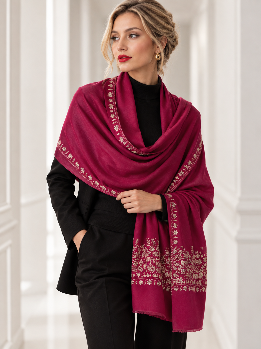 A woman wearing a black outfit and a deep magenta shawl with intricate gold floral borders, standing in a bright white hallway.