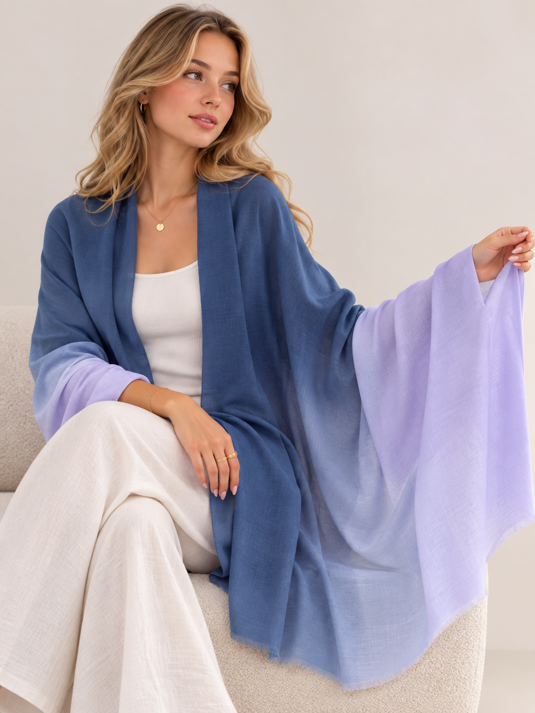 A woman seated indoors wearing a lightweight open-front shawl draped over her shoulders. The shawl has a blue-to-lavender ombré color gradient and is shown against a soft neutral interior background.