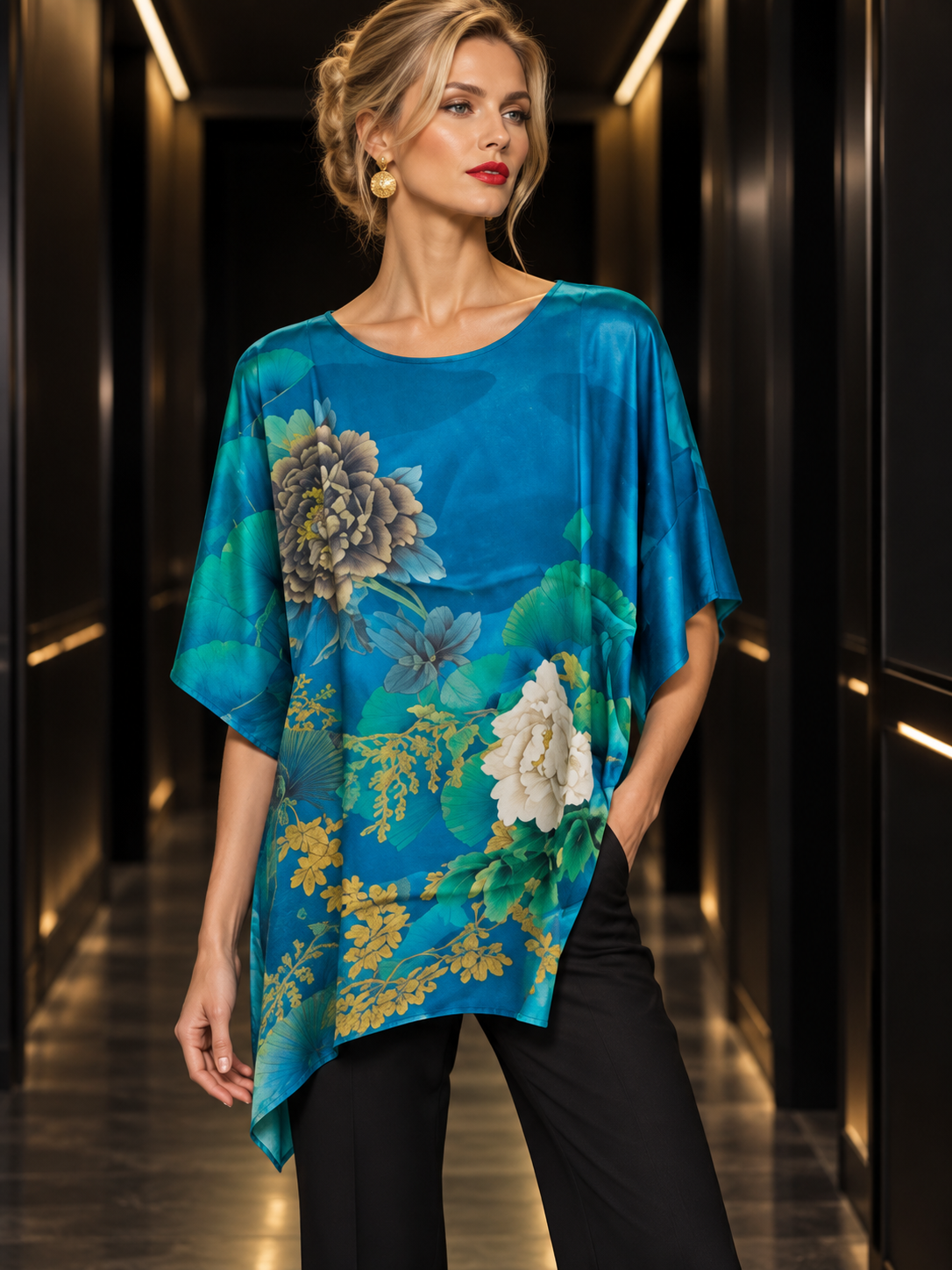 A woman models a vibrant blue silk top with a large floral print in green, white, and yellow, paired with black pants, standing in a dark hallway.