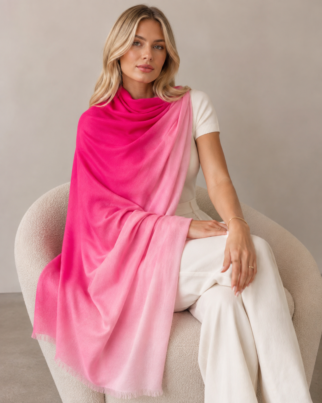 A woman seated on a light beige armchair wearing a large gradient shawl scarf that fades from bright pink at the top to soft light pink at the bottom, paired with a white top and white pants.