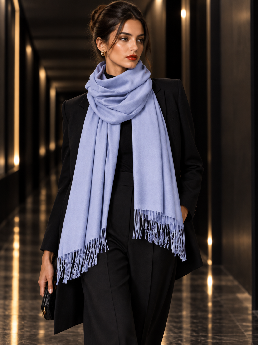 A woman wearing a light periwinkle blue scarf with fringed ends, styled over a black blazer and trousers, walking down a modern, dimly lit hallway.
