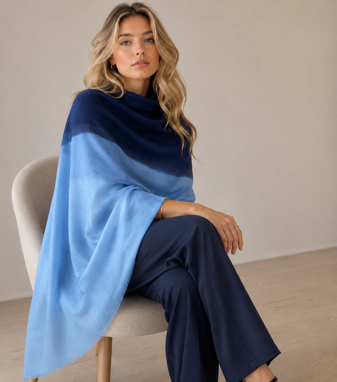 A woman sits on a light beige chair wearing a large draped shawl in two shades of blue, with navy at the top blending into light blue below. She is also wearing navy wide-leg pants in a minimalist indoor setting.