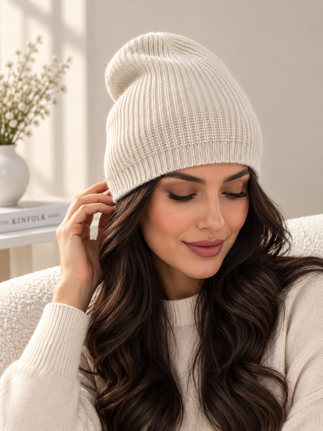 A woman wearing a cream-colored ribbed knit beanie hat and a matching cream sweater, sitting in a cozy indoor setting with soft natural light.