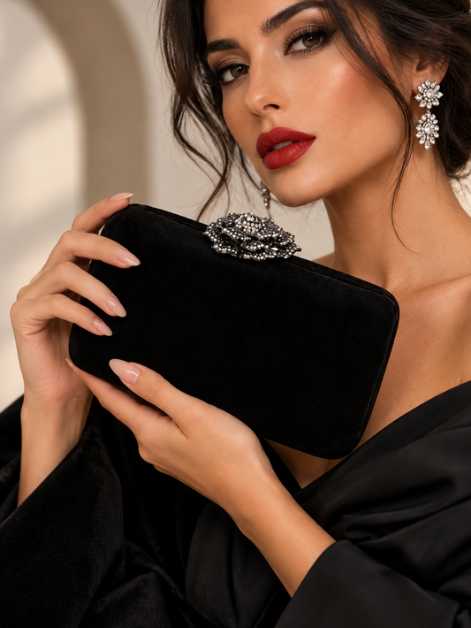 A woman in a black satin dress holds a black velvet clutch decorated with a sparkling silver crystal rose clasp. She is wearing matching crystal flower drop earrings and has dark hair and red lipstick.