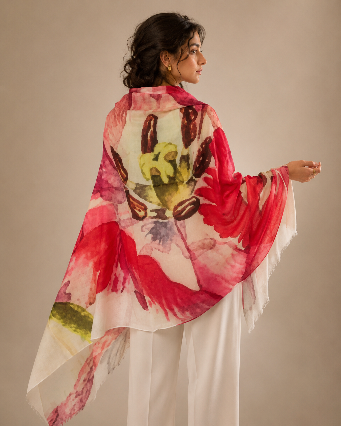 A woman shown from the back wearing a lightweight draped shawl or wrap with a large watercolor-style floral print in red, pink, cream, and olive green, paired with white pants against a plain beige studio background.