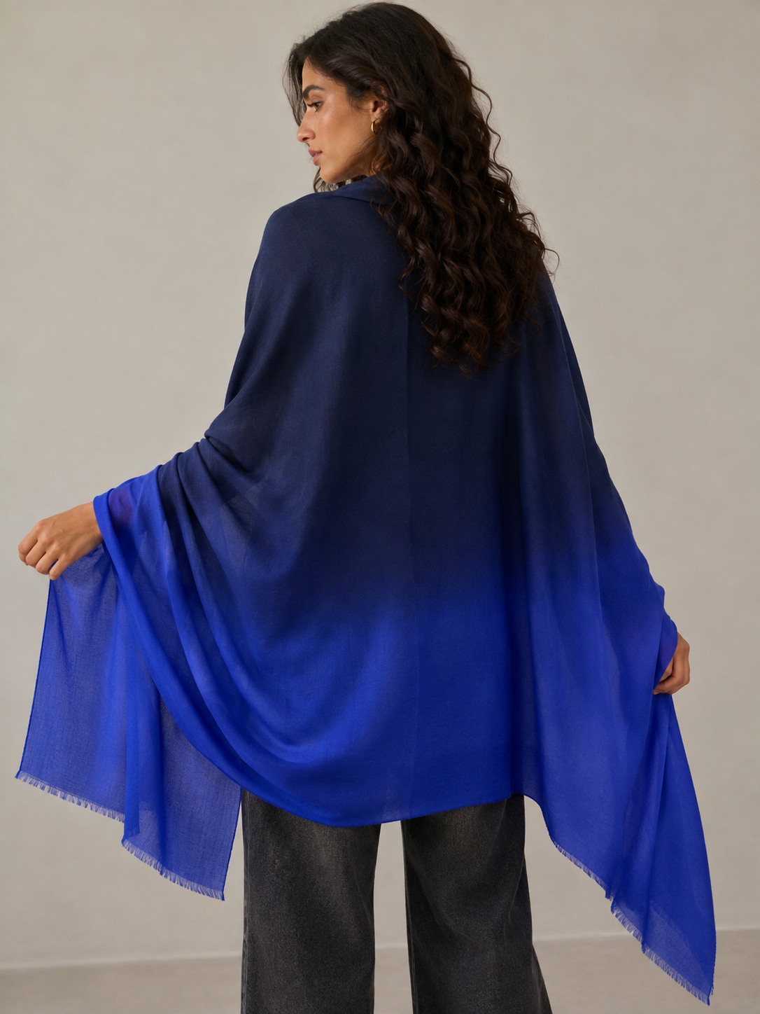 A woman wearing a large blue ombre shawl or poncho draped over her shoulders, shown from the back against a plain light gray studio background.
