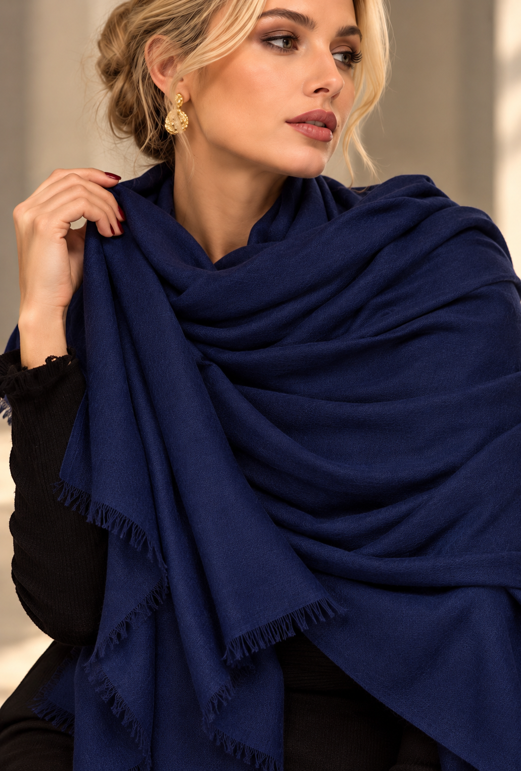 A woman wearing a large navy blue shawl with fringed edges, draped over her shoulders and held in place by a black top. She is wearing gold earrings, and the background is a softly blurred neutral indoor setting.