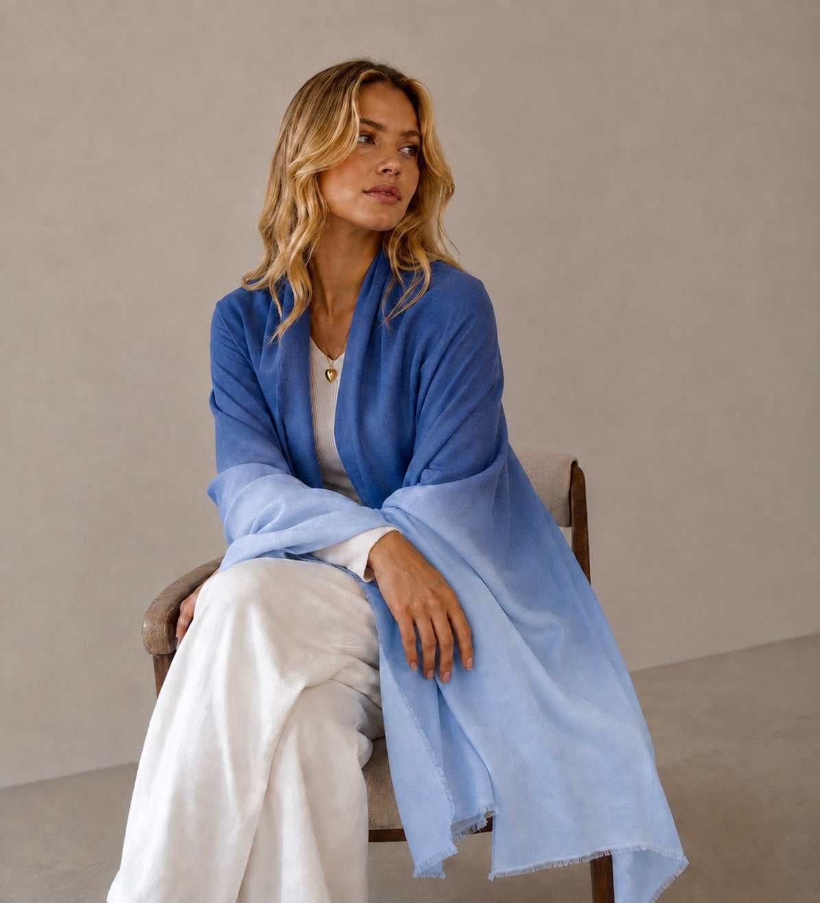 A woman sits on a wooden chair wearing a light blue gradient shawl draped over her shoulders, styled over a white top and white trousers, against a neutral beige background.
