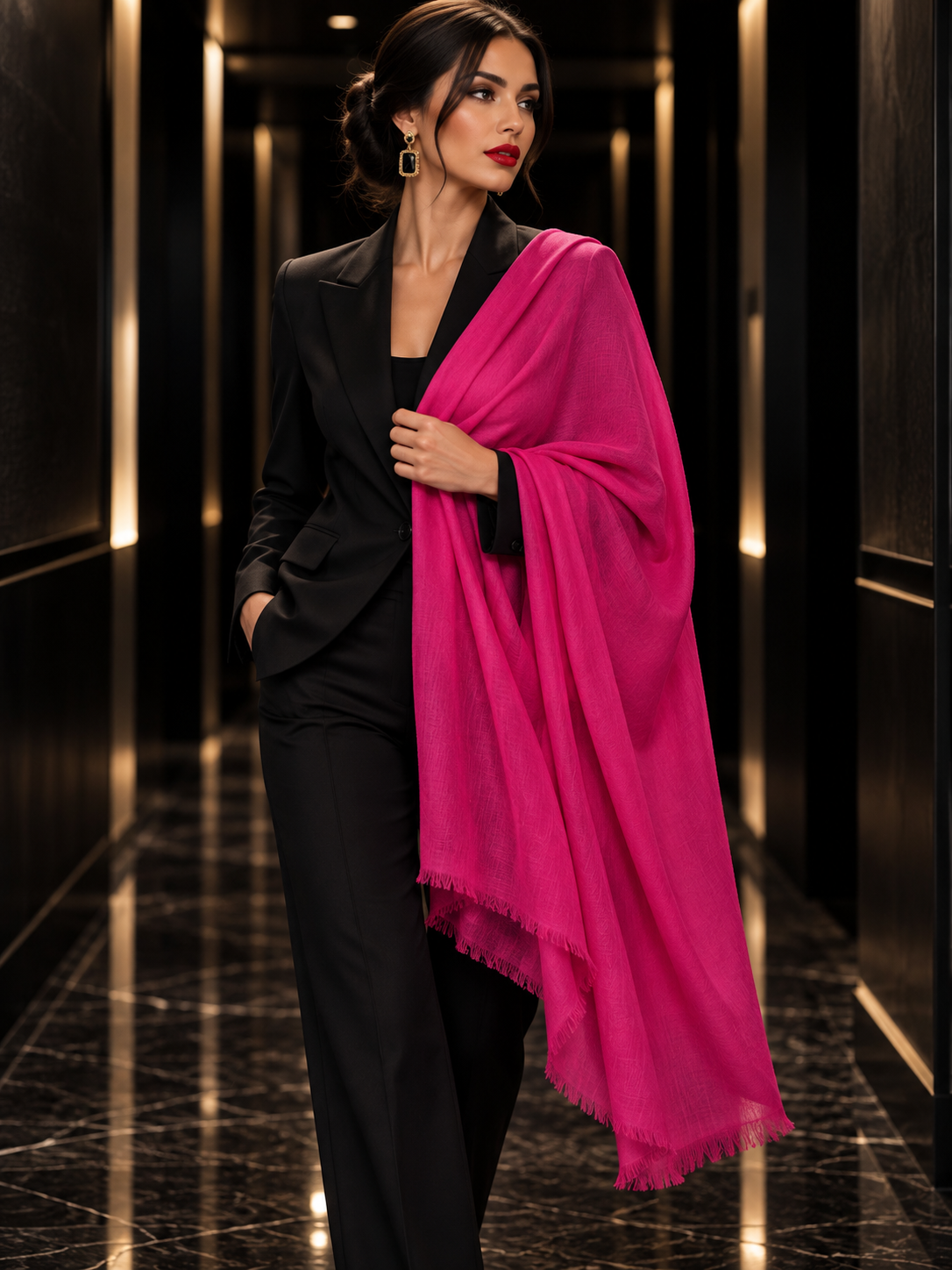 A woman in a black pantsuit stands in a dark hallway, draped with a bright pink shawl or scarf with fringed edges.