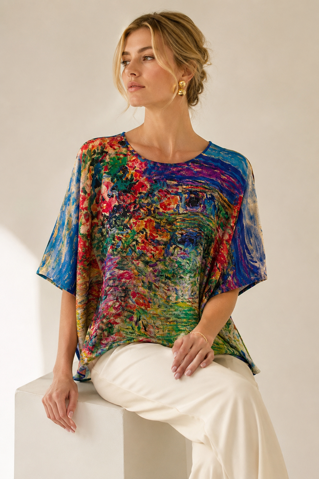 "Printed silk tunic with Impressionist garden floral print in cobalt blue, red, and green — relaxed batwing silhouette, seated editorial shot"