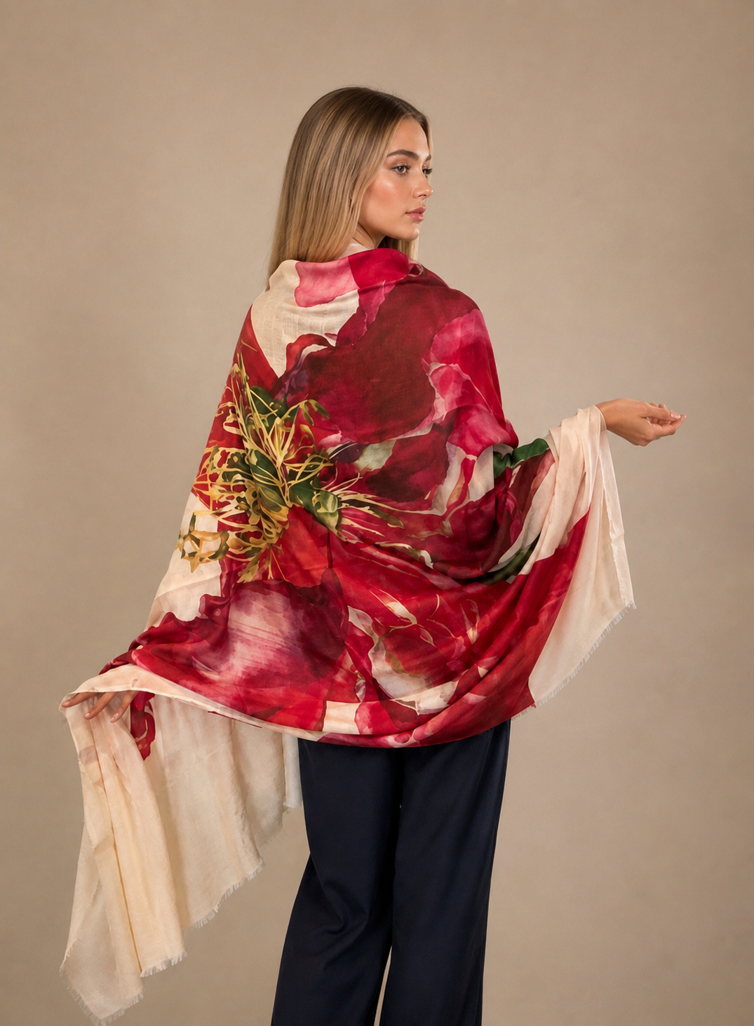 A woman wearing a large floral shawl wrap in deep red, pink, and cream with green leaf accents, paired with dark navy pants, posed against a solid beige studio background.