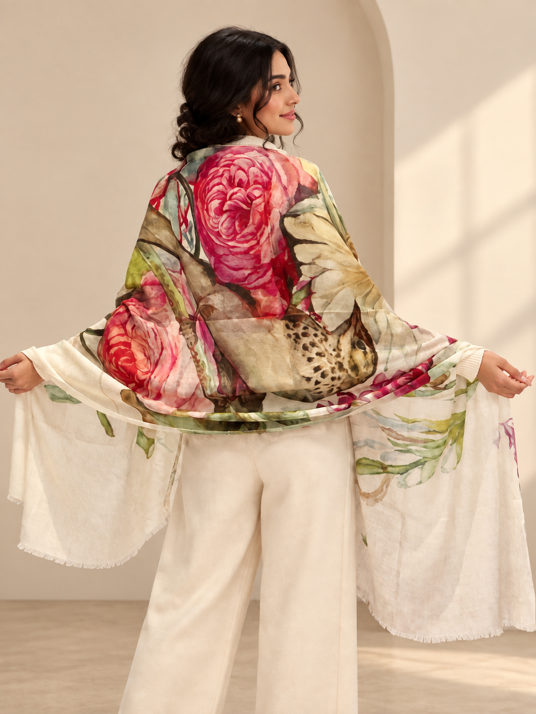 A woman wearing a white wide-leg pants set and a large floral shawl draped over her back, shown in a warm indoor setting with soft sunlight and shadows.