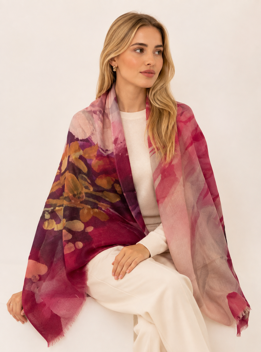 A woman seated against a plain light background wearing a large semi-sheer rectangular scarf with a watercolor floral pattern in pink, magenta, and gold tones.